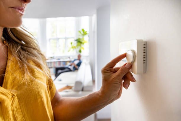 Woman in yellow shirt adjusting a white thermostat on a wall indoors.