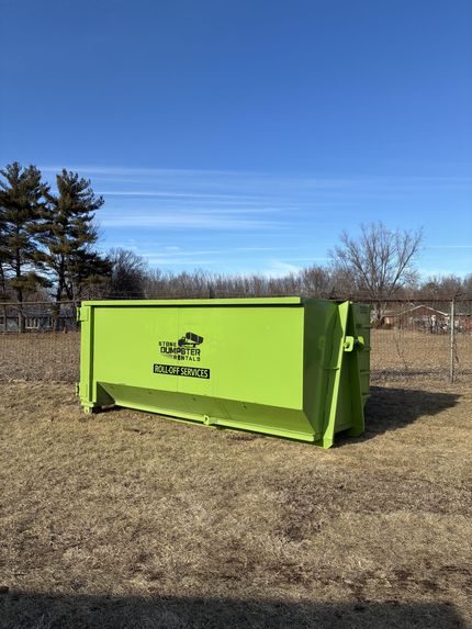 A large, brown, rusted roll-off dumpster filled with various construction debris, parked on a concrete path near grass. A large, brown, rusted roll-off dumpster filled with various construction debris, parked on a concrete path near grass.
