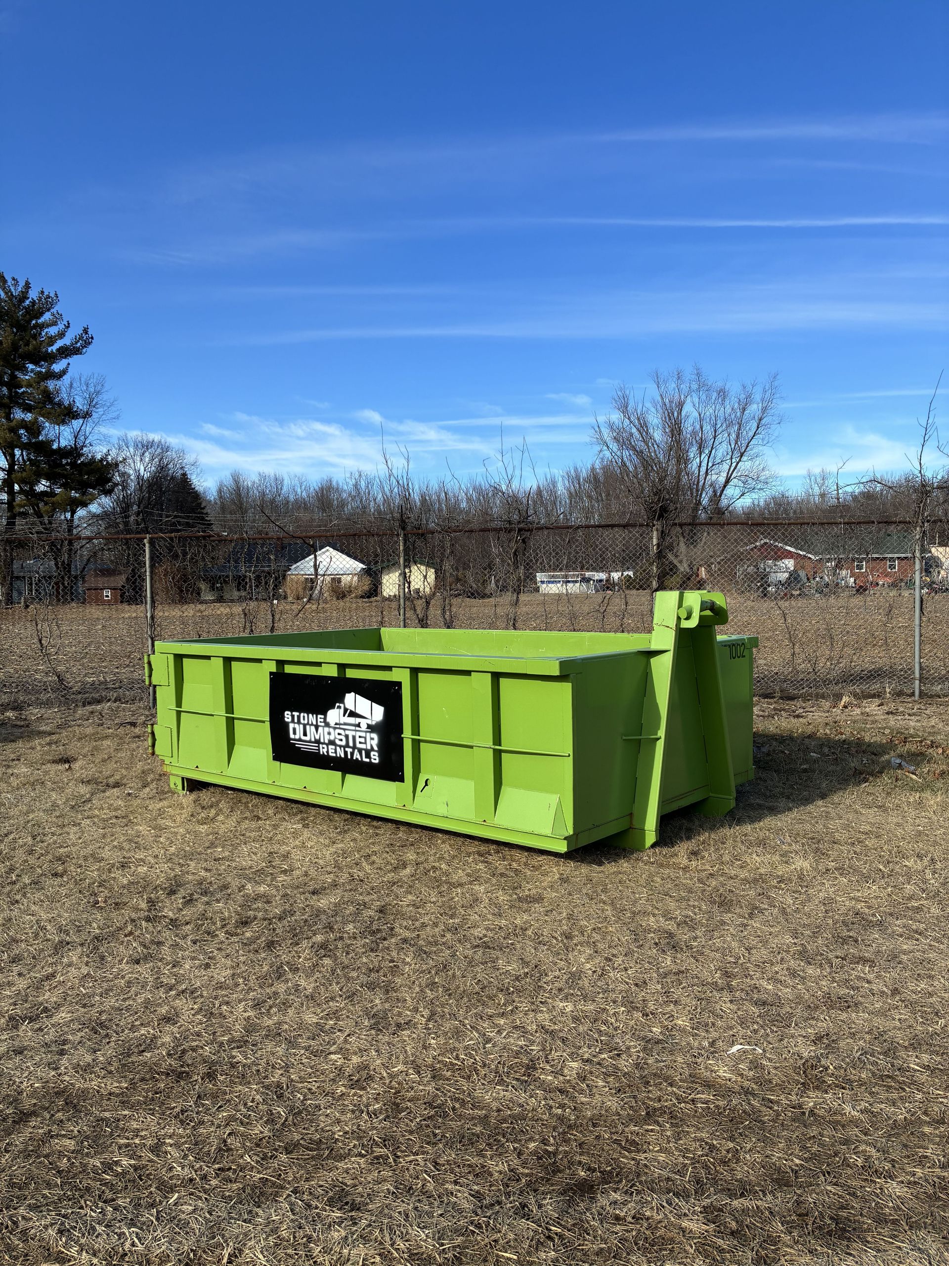 A large black metal dumpster sits on a paved driveway in front of a brick house with a front porch.