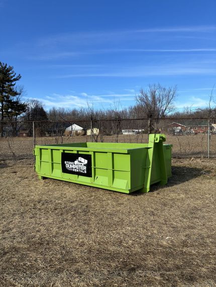 A blue metal dumpster sits on a paved driveway in front of a white house with trees in the yard. A blue metal dumpster sits on a paved driveway in front of a white house with trees in the yard.