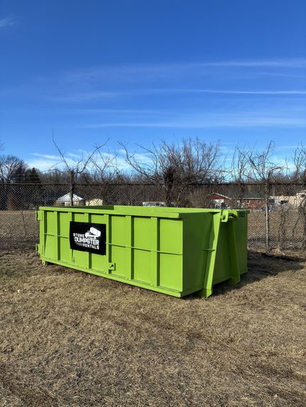 A large, empty blue dumpster sits on a dirt lot in front of a modern residential home under construction. A large, empty blue dumpster sits on a dirt lot in front of a modern residential home under construction.