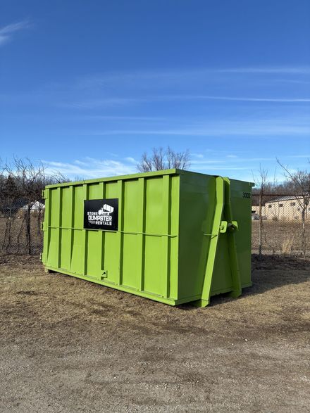 A large, empty green roll-off dumpster sitting on a paved surface with bare trees in the background. A large, empty green roll-off dumpster sitting on a paved surface with bare trees in the background.