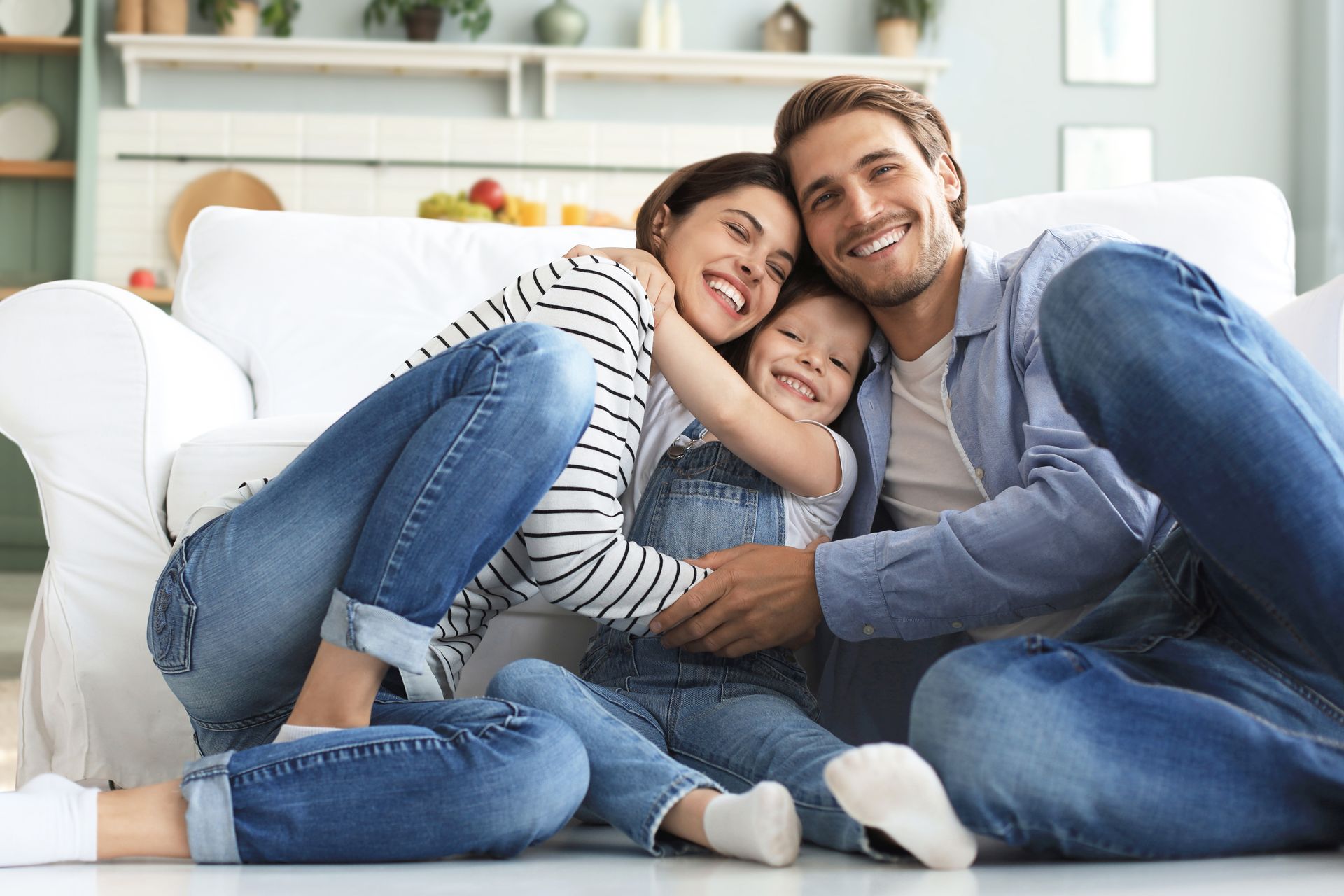 Family of four on a bed, embracing. Parents kiss, holding two young children. White bedding and walls.