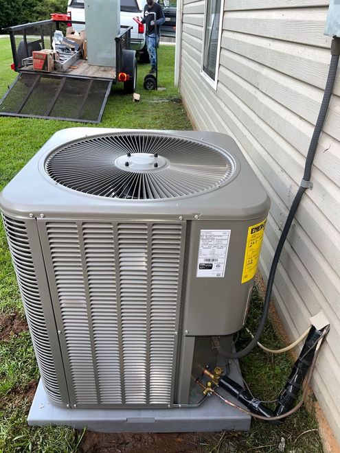 A new gray residential air conditioning unit sits on a concrete pad next to a house, with a utility trailer in the grass.