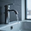 Rubinetto cromato con acqua che scorre in un lavandino bianco. Sfondo piastrellato scuro, finestra luminosa sulla destra.