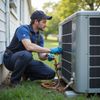 Un tecnico HVAC si inginocchia all'esterno e, indossando i guanti, esamina l'unità AC.