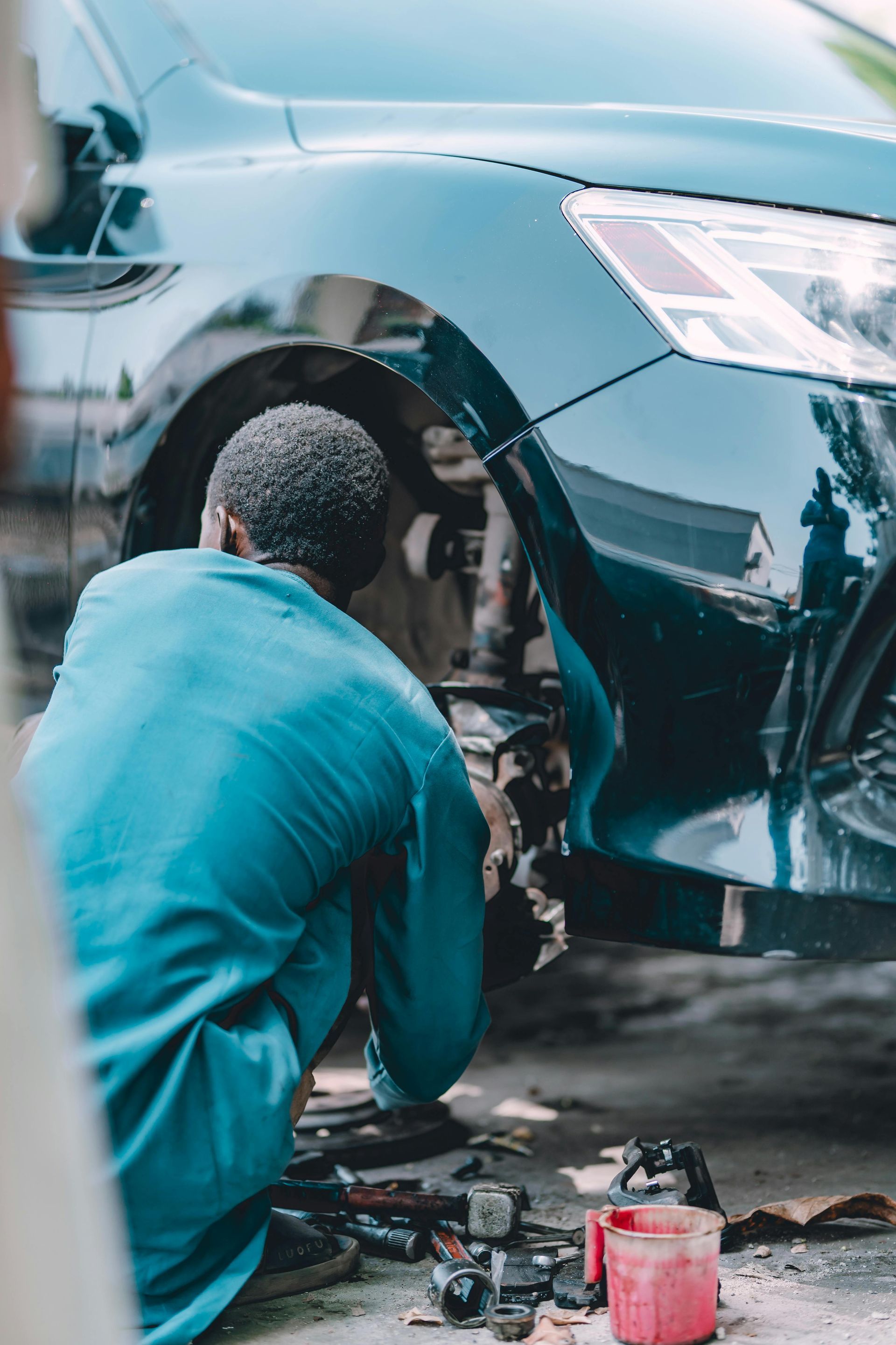 A mechanic in a blue uniform works on the wheel assembly of a dark green car, with tools scattered on the ground.