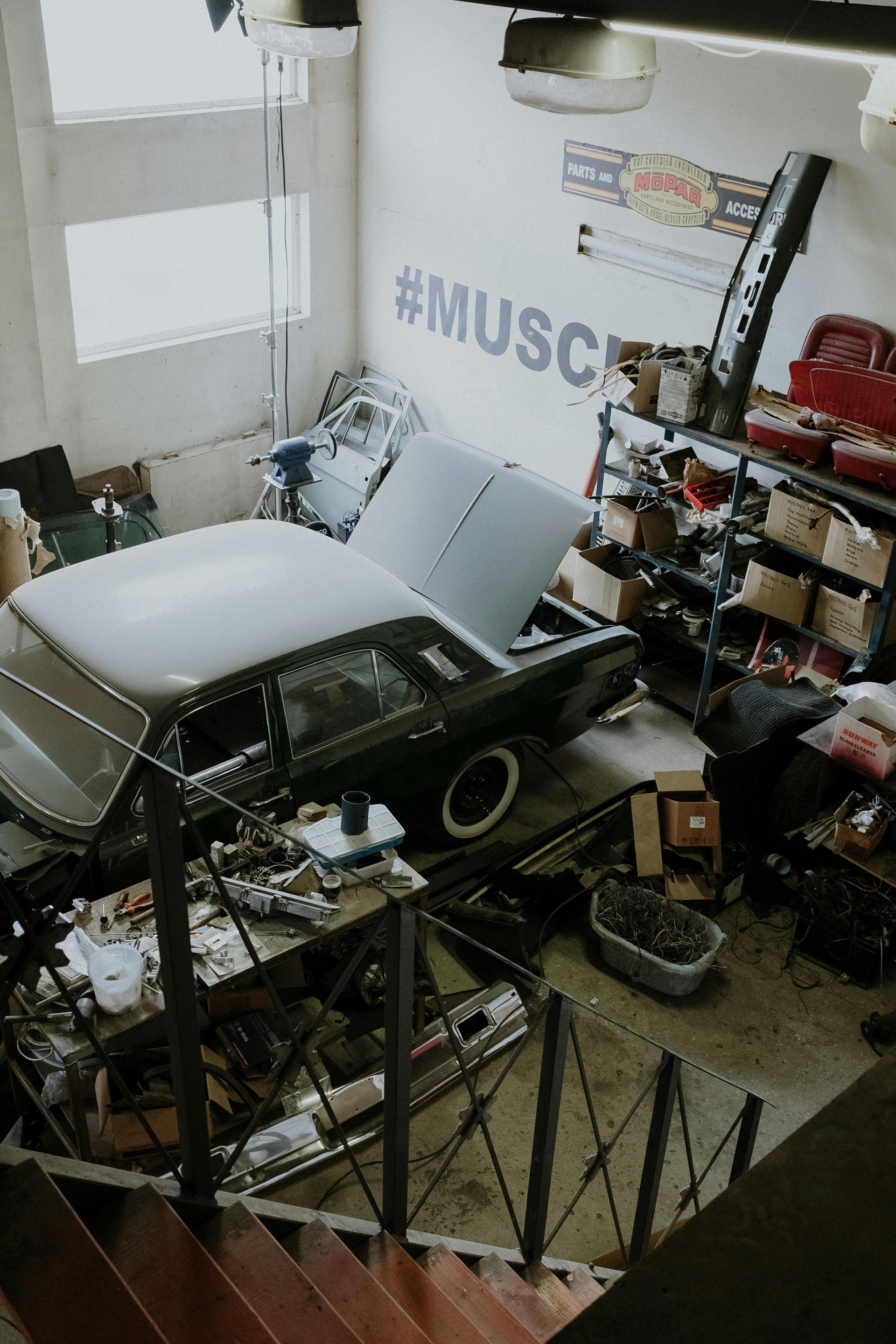An elevated view of a cluttered car workshop featuring a dark vintage car, shelving units, and a staircase.