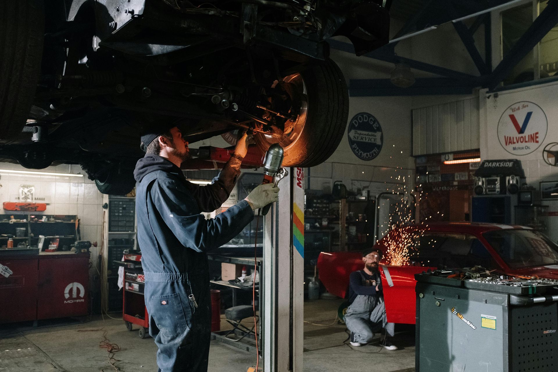 A mechanic uses a power tool on a car raised on a lift, while another person works on a red car nearby in a garage.