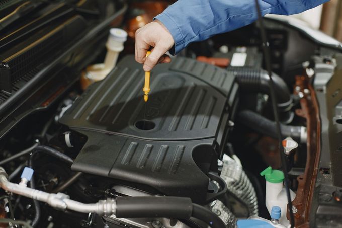 A mechanic in a blue uniform checks the engine oil level with a dipstick inside a car's engine bay.