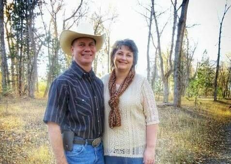 a man and a woman are posing for a picture in the woods . the man is wearing a cowboy hat .