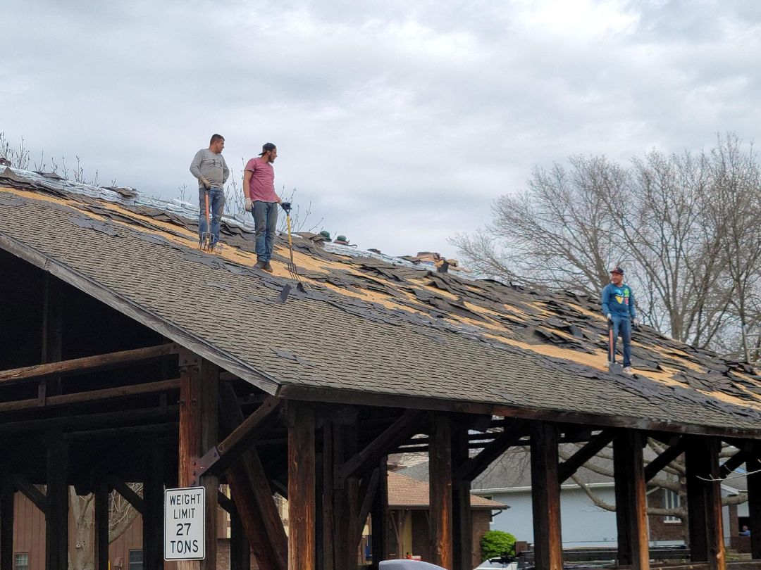 Three people removing shingles from a wooden roof of a pavilion under an overcast sky.