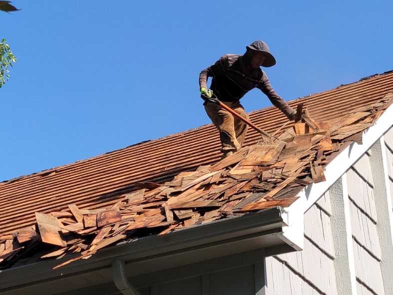 Person on a roof removing old shingles with a tool, against a blue sky.