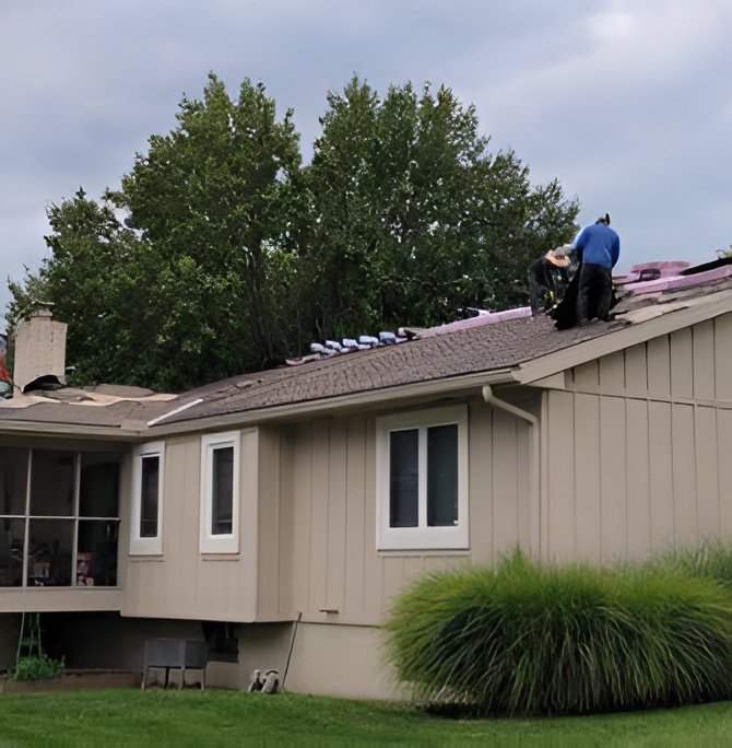 Roofers working on a residential roof, some shingles removed, cloudy sky, green grass.
