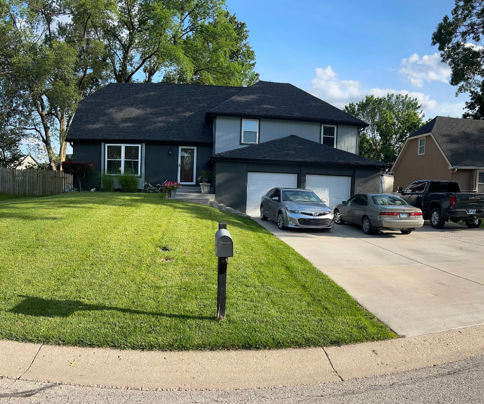 Two-story house with gray facade, black roof, and manicured lawn. Two cars parked in the driveway.