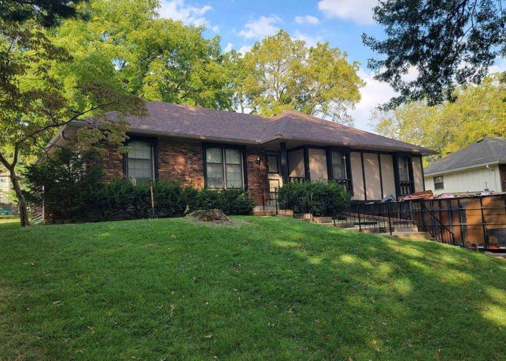 Low brick house with brown roof and green lawn, trees in the background.