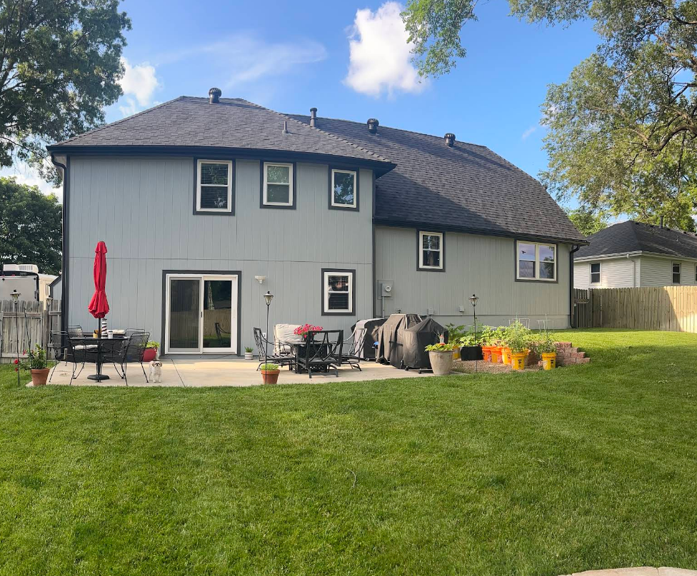 Backyard view of a two-story gray house with patio, garden, and green lawn on a sunny day.