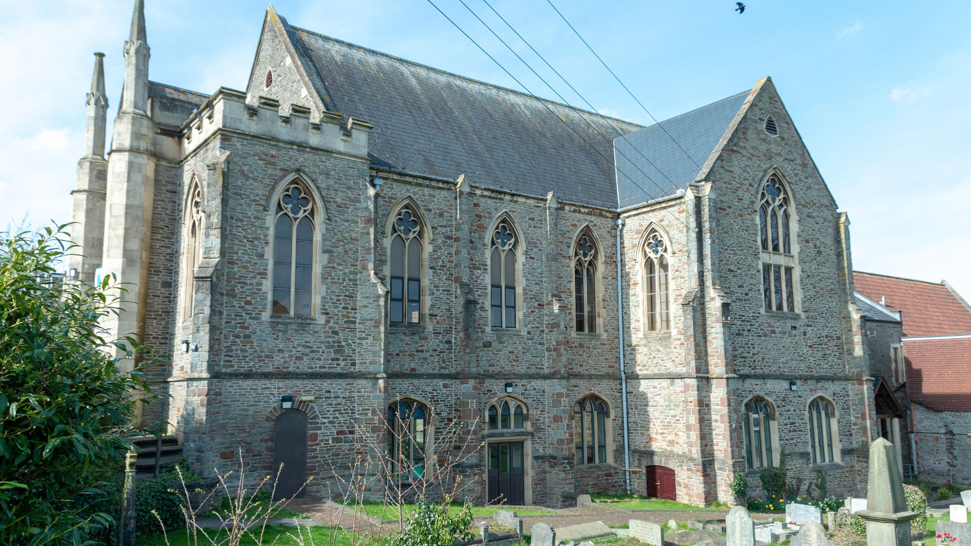 Gothic style stone church with arched windows and a gray slate roof, in front of a graveyard.