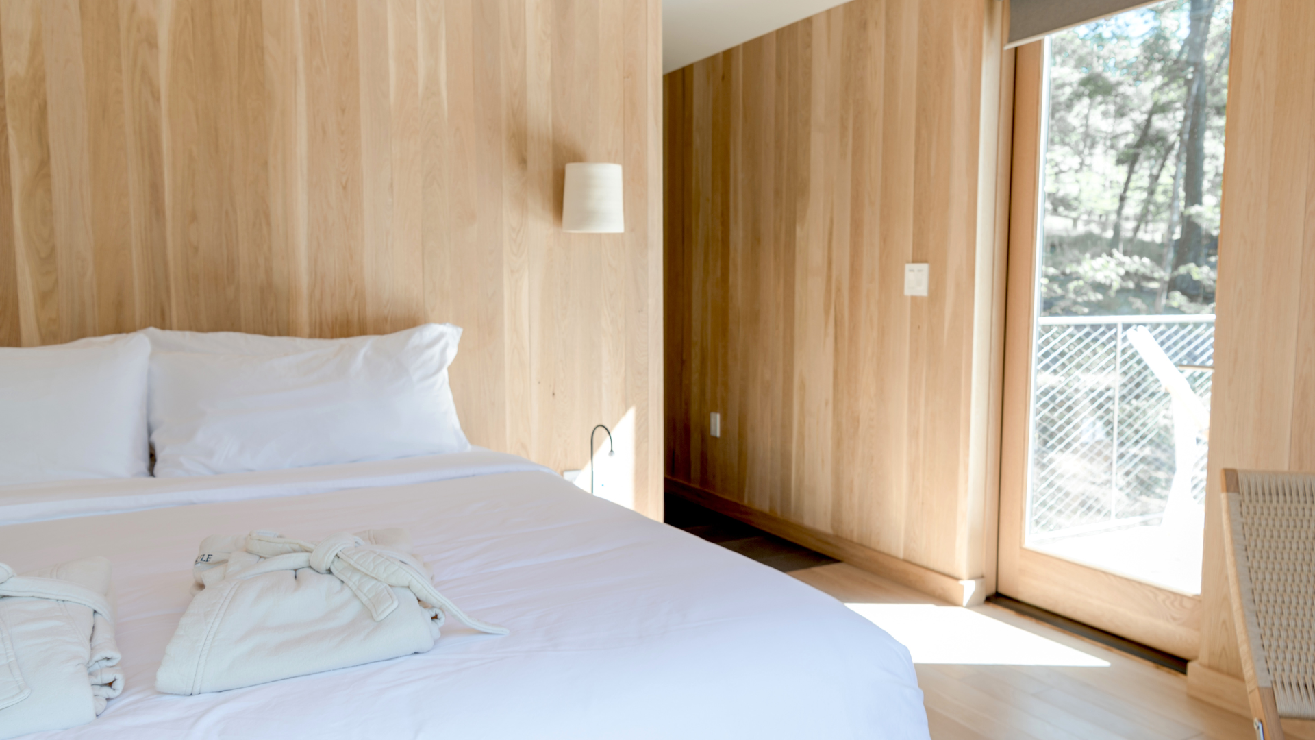 Bedroom with light wood paneling, white bedding, and a glass door leading to a balcony with tree views.