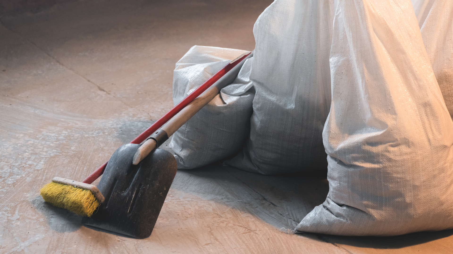 Shovel and broom leaning against two large white bags on a concrete floor.