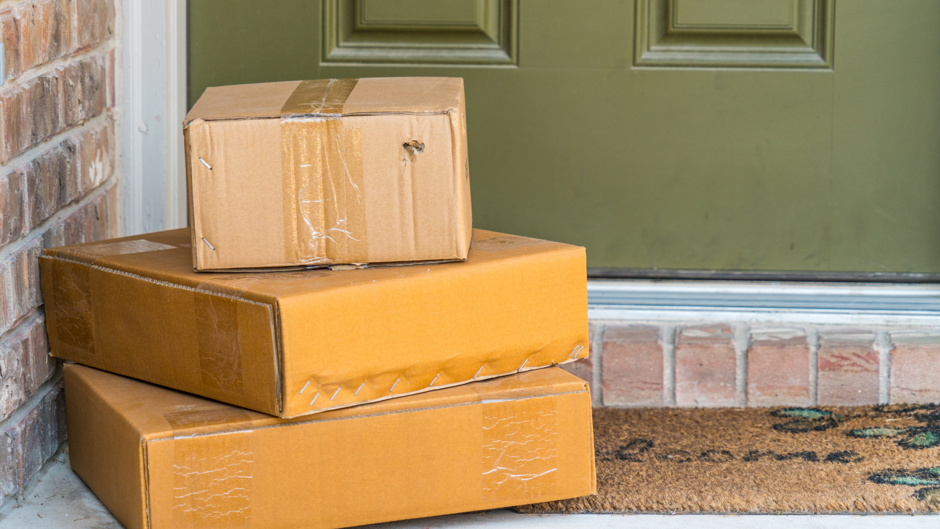 Three brown cardboard packages stacked in front of a green door with a welcome mat on a brick porch.