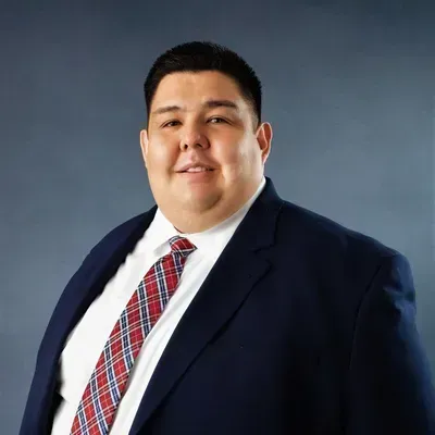 Man in navy suit, white shirt, and red-plaid tie smiles, chest-up, against a blue background.