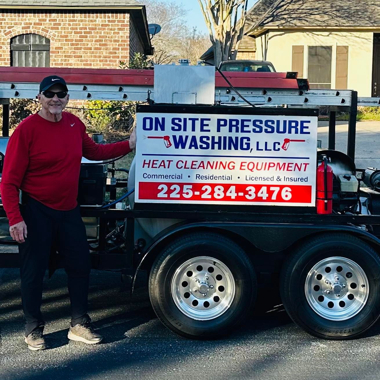 Man in red shirt stands by a pressure washing trailer, holding sign with company name and phone number.