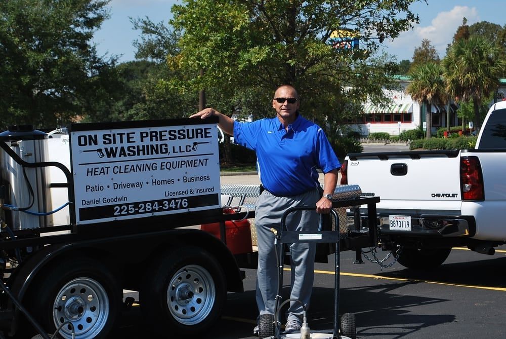 Man standing with pressure washing equipment trailer. Blue shirt, gray pants, sunny day.
