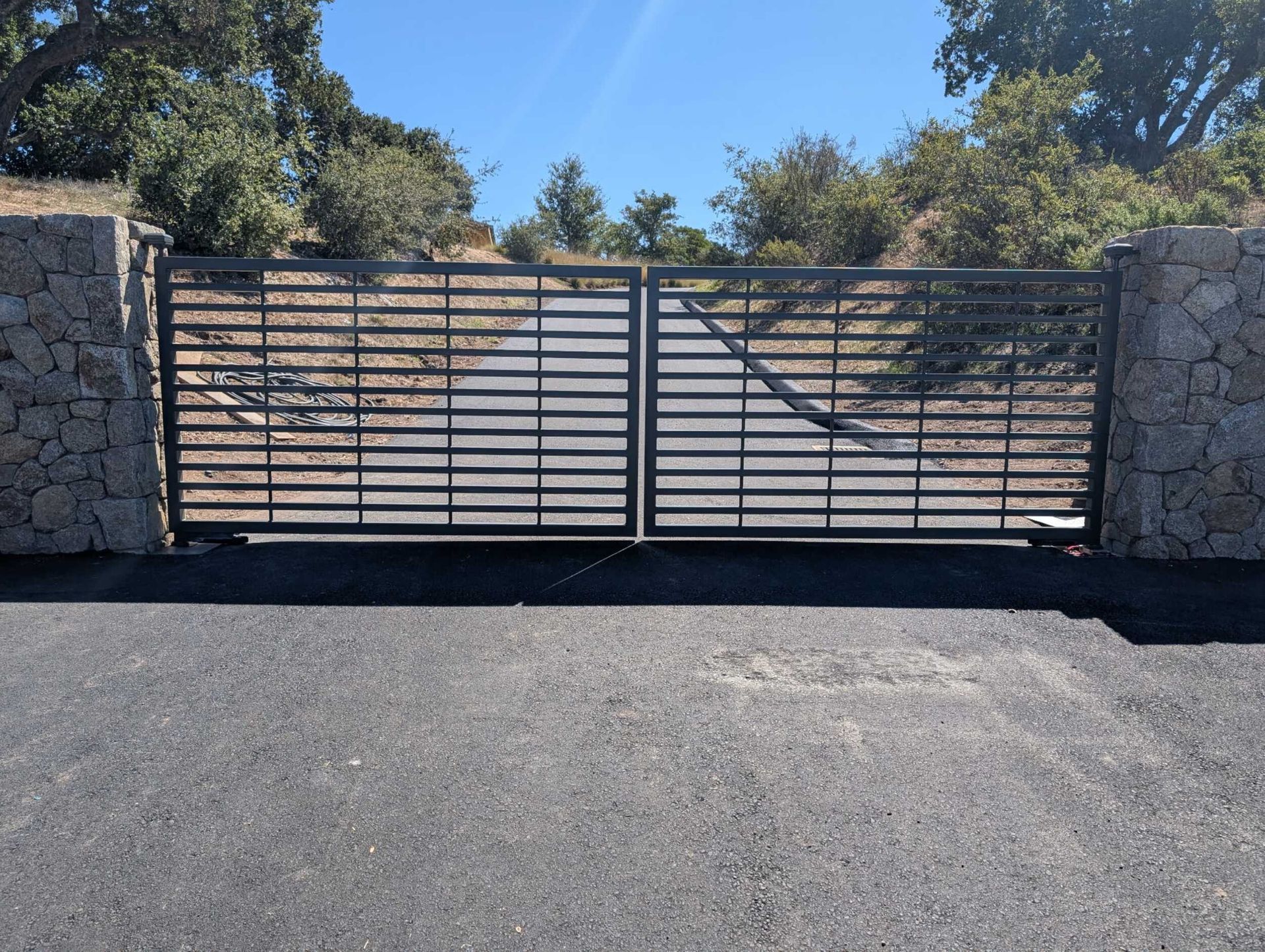 Black metal gate set between stone pillars on a paved road, leading toward trees.