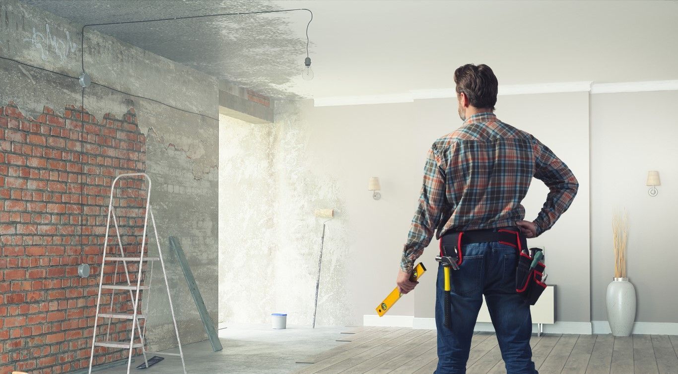 Worker holding a drill in a partially renovated room with exposed brick, scaffolding, and paint buckets