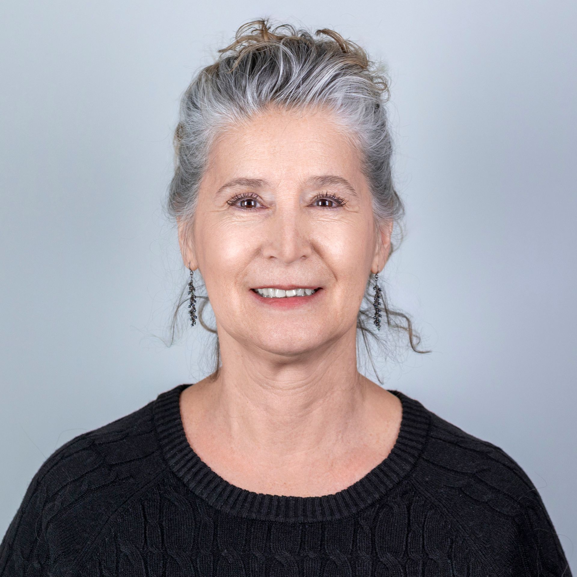 Woman with gray hair smiles, wearing a black sweater, against a light gray background.