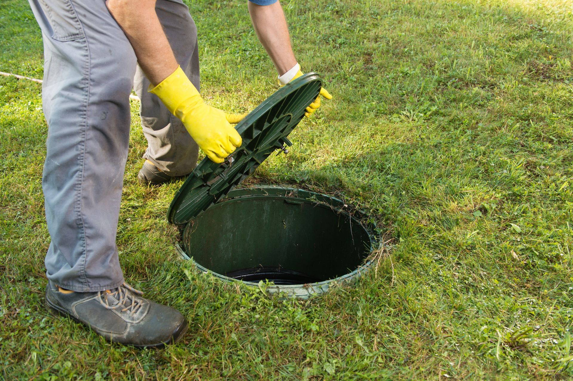 Worker lifting a septic tank lid in a grassy yard.