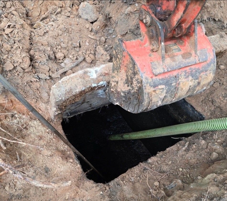 An excavator bucket unearths a dark pit, with a green hose extended into it.