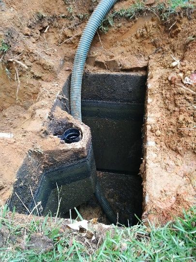 Open utility pit in dirt with a gray corrugated hose and black lining beside grass