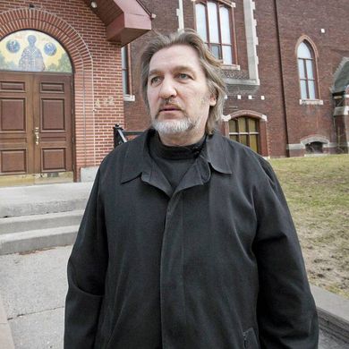 A man in a black jacket stands in front of a brick building