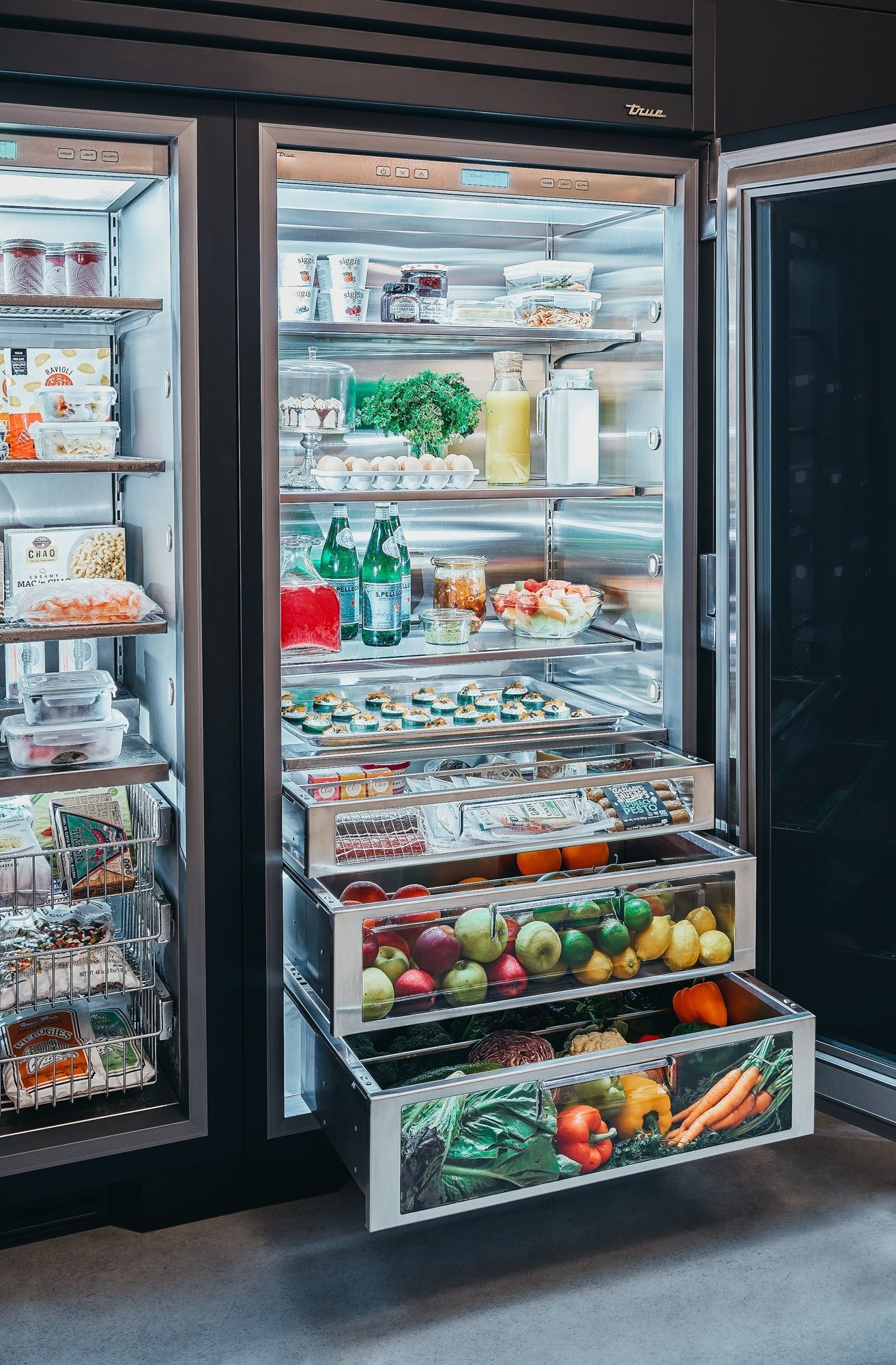 Open refrigerator showcasing fresh produce and pre-packaged food. Drawers and shelves are illuminated with bright lighting.