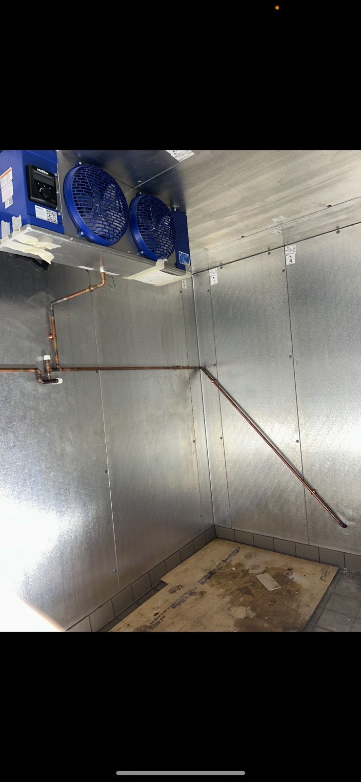 Interior of a walk-in cooler with silver insulated walls, copper piping, and a wooden floor panel.