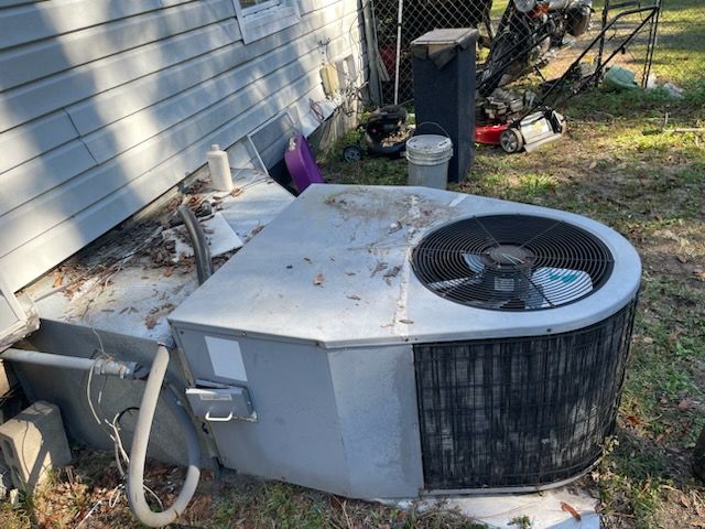 An outside air conditioner unit, gray and dusty, next to a house with debris nearby.
