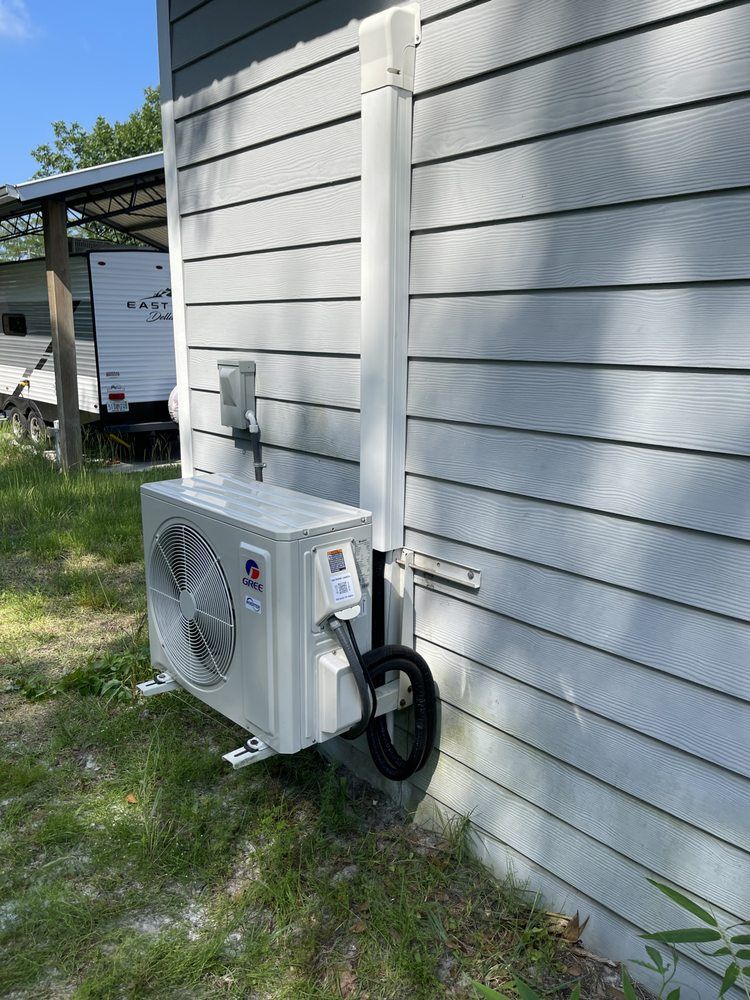 An outdoor air conditioning unit mounted on a gray-sided house with a white conduit running up the wall.
