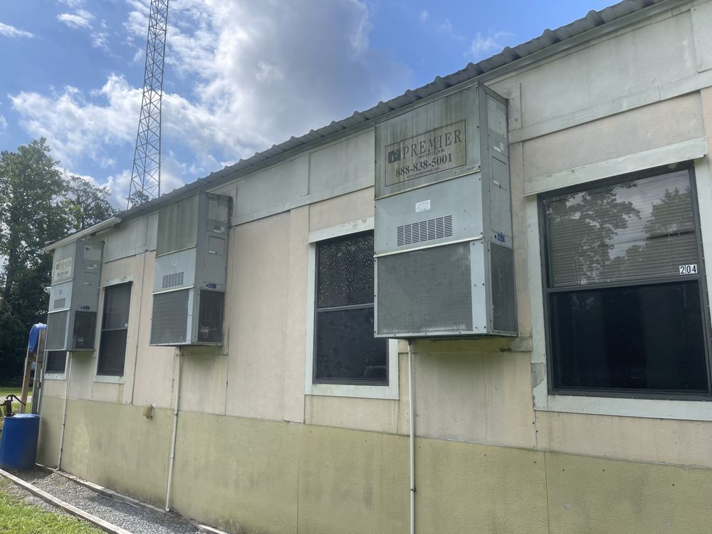Exterior of a building with multiple window air conditioners, under a metal roof, and a cellular tower in the background.