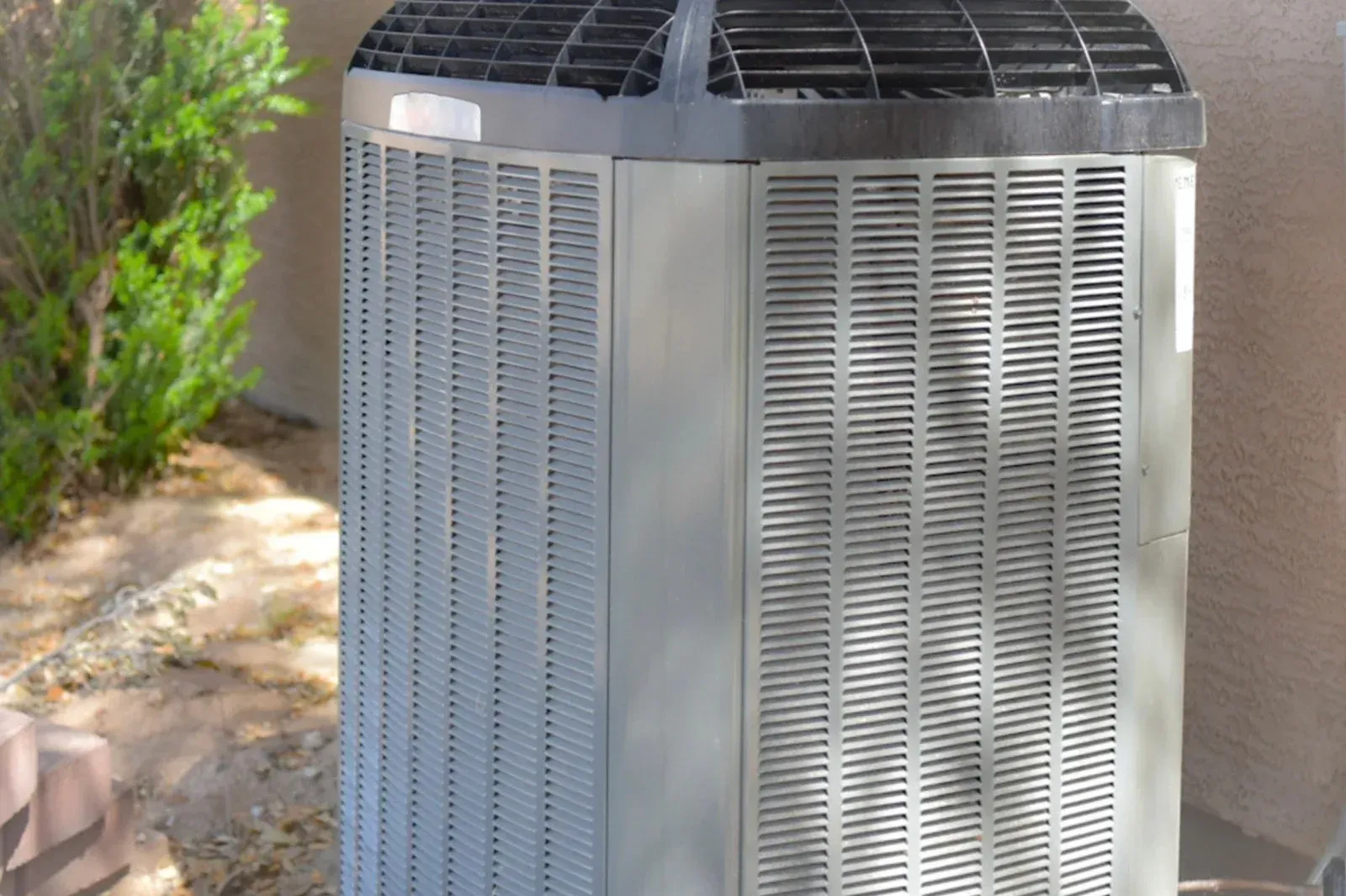 Air conditioning unit outside a building, gray with dark top, near a bush.