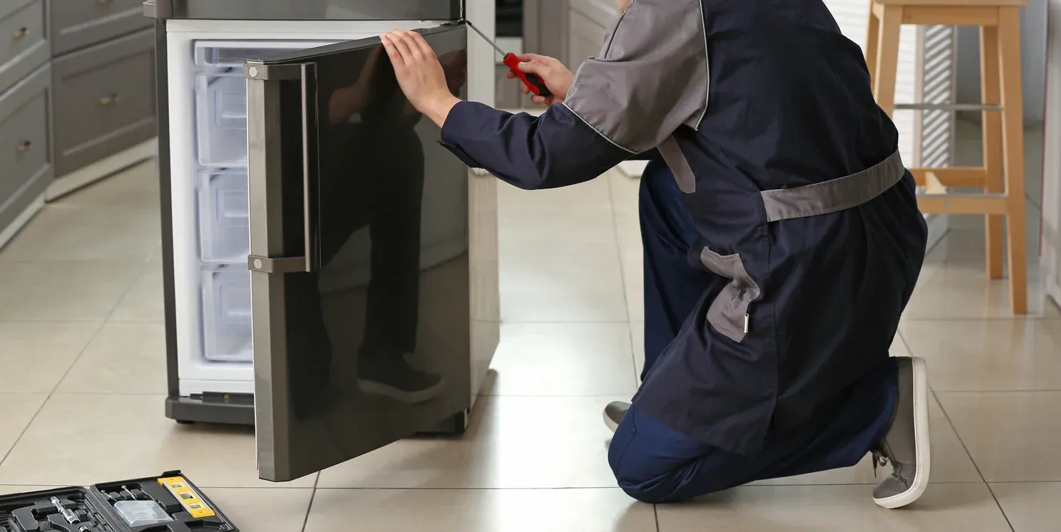 A person in a blue jumpsuit repairs a refrigerator, holding a screwdriver.