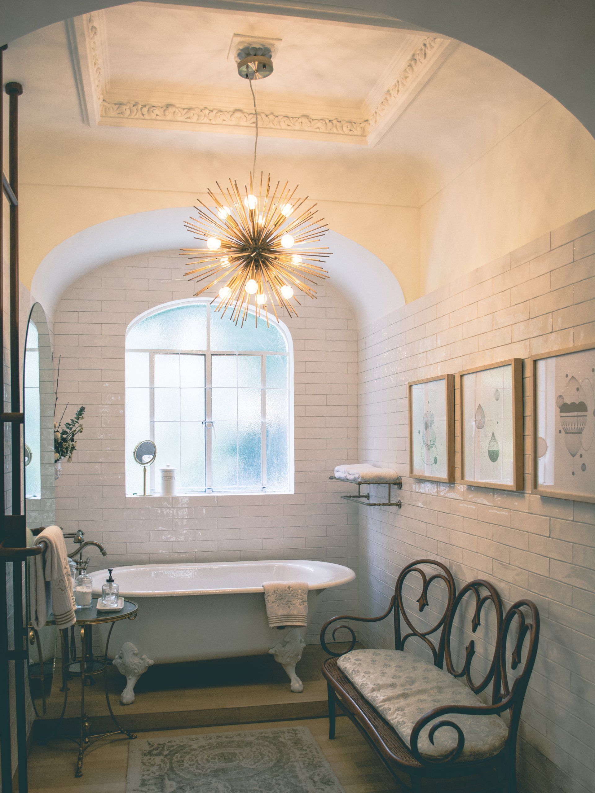 An old style bathroom with a tub , bench and chandelier.