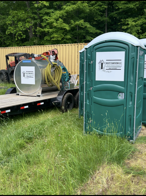 Green portable toilets next to a trailer with a tank and equipment; in grassy area.
