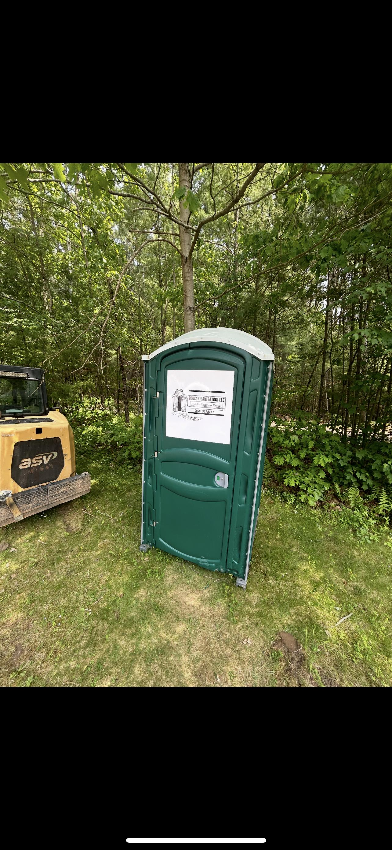 Green porta-potty in a grassy area, partially obscured by trees. A small construction vehicle is on the left.