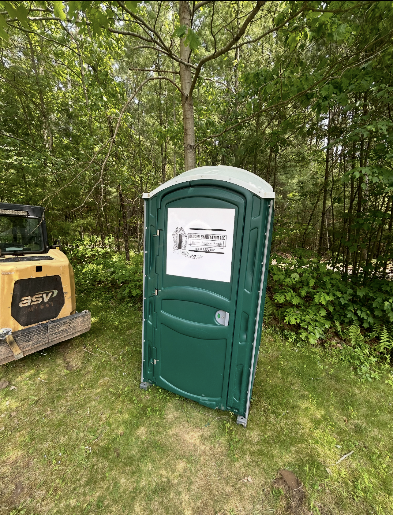 Trailer with a water tank and hose next to a porta-potty in a wooded area.