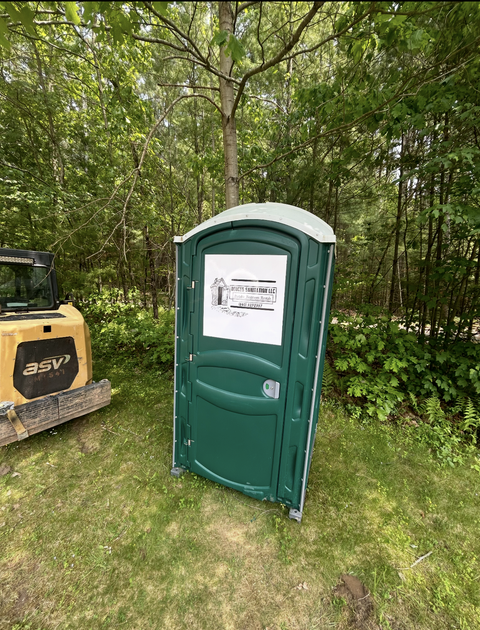 Trailer with a water tank and hose next to a porta-potty in a wooded area.