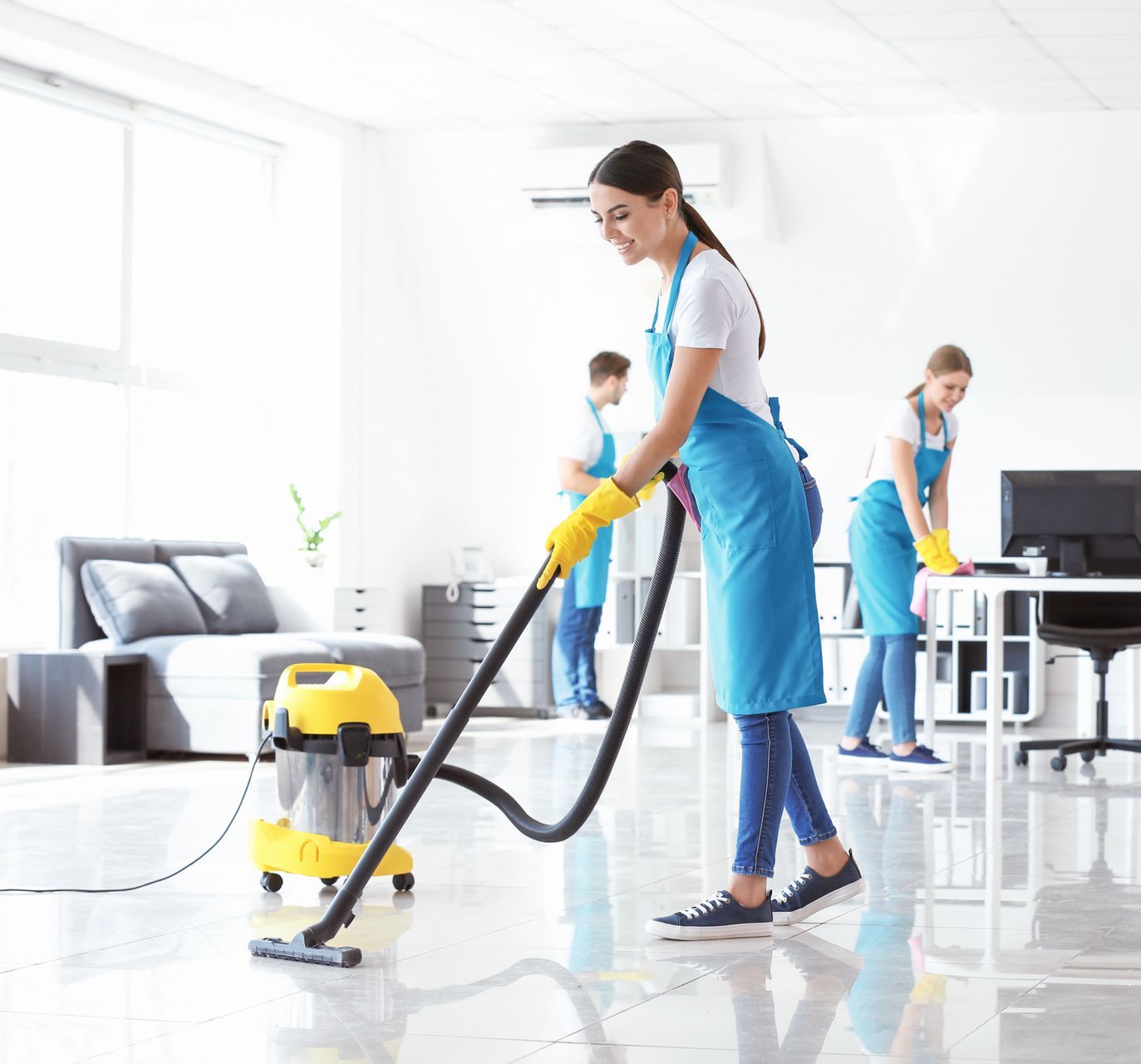 Une femme passe l'aspirateur dans un bureau, avec deux autres agents d'entretien en arrière-plan. Ils portent des tabliers bleus.