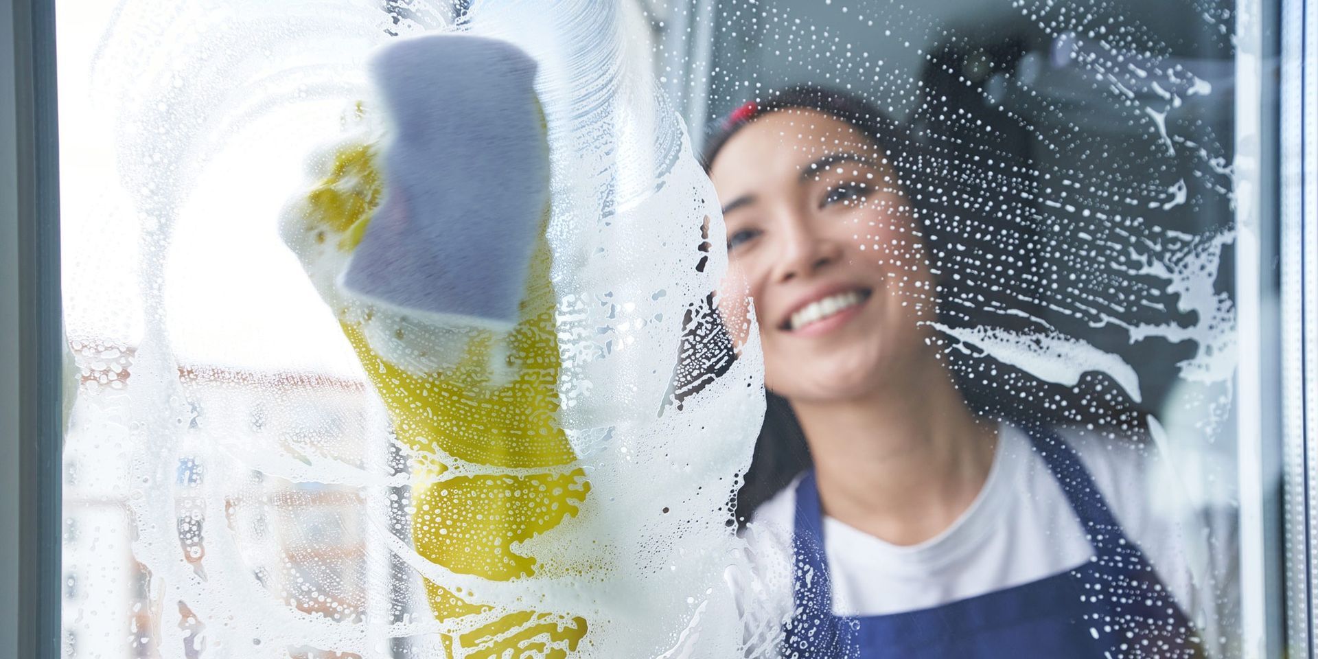 Une femme nettoie une fenêtre avec une éponge et sourit.