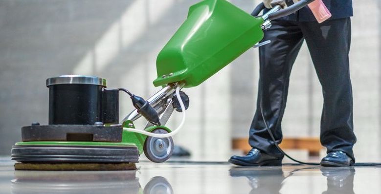 Une personne en pantalon et chaussures noires utilise une polisseuse de sol verte pour nettoyer un sol gris clair brillant.